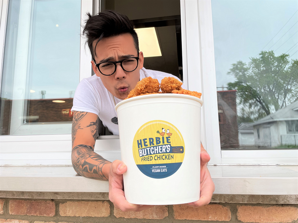 a person hanging out of a takeout window holding a big bucket of vegan fried chicken