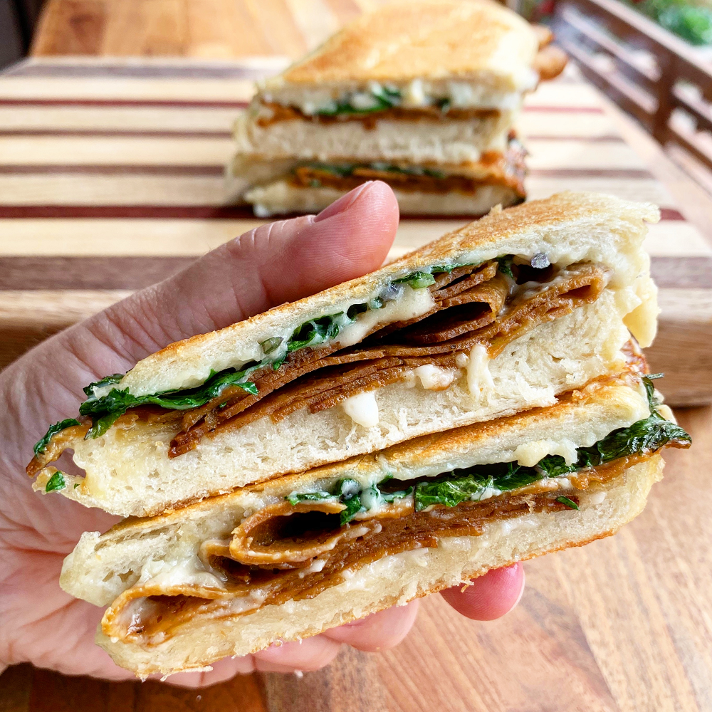 hand holding two halves of a vegan sandwich with plant based roast beef, dairy free cheese and greens